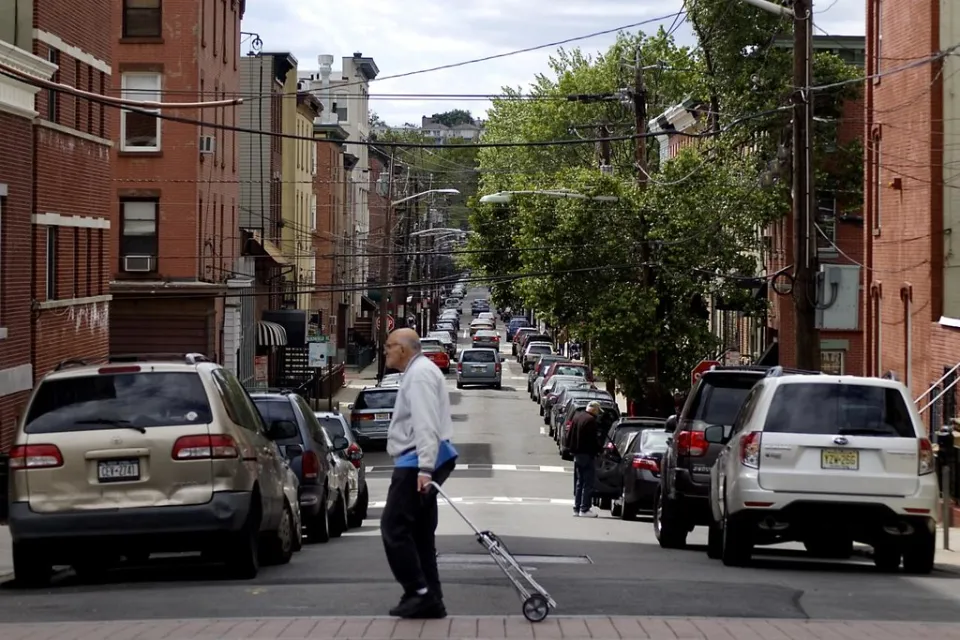 An old man crosses a one-way street in the foreground, with both sides of the streets lined with cars facing away from the camera and brick buildings.